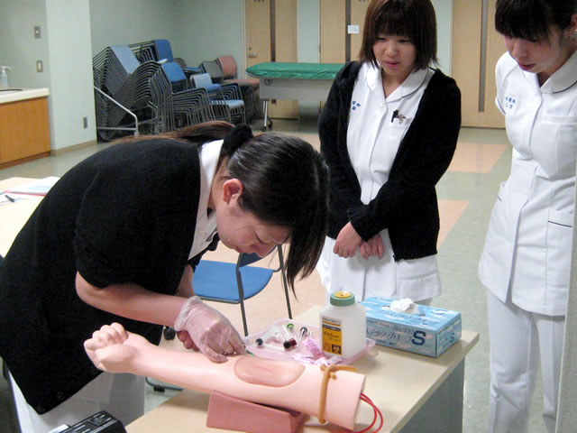 左腕のみの研修用人形で採決法の研修を行っている女性看護師とそれを見ている二人の女性看護師