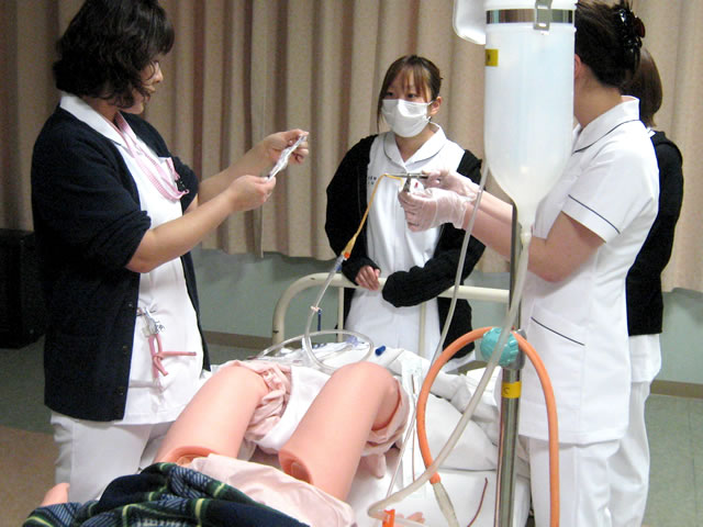 研修用人形で導尿・留置カテーテル挿入法の研修を行う女性看護師と話を聞く女性看護師たち