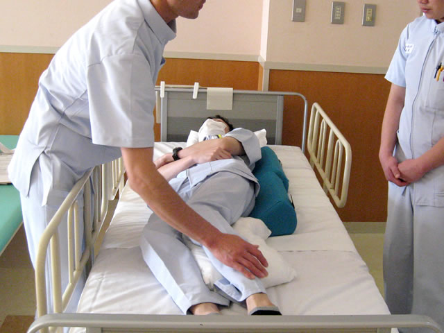 ベッドに寝ている男性で体位の調整の研修を行う男性看護師と話を聞く女性看護師